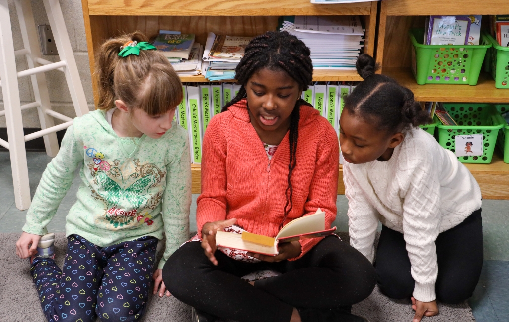 Three students, one in the middle reads to the two students sitting on side of them