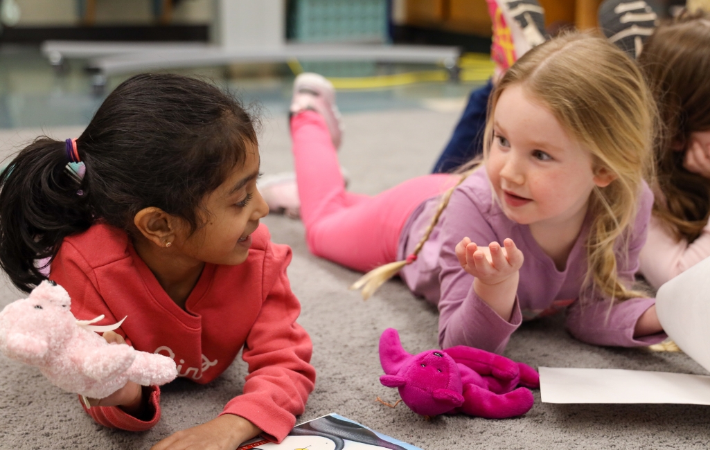 Two students chat while laying on the rug reading, holding stuff animal reading buddies.