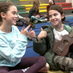 Two students give each other a thumbs up, while one holds a ukulele in music class.