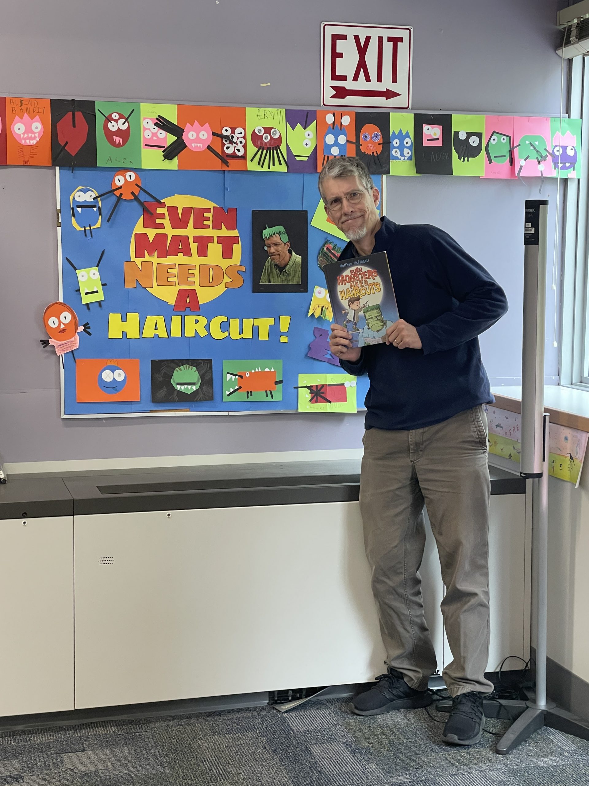 Photo of Matt McElligott next to a display in the library that says" Even Matt Needs a Haircut", a play on one of his book titles