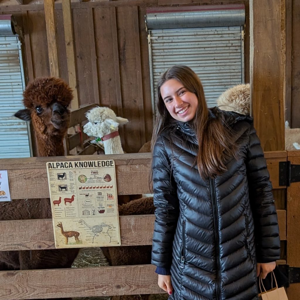 student poses for photo while alpaca looks on